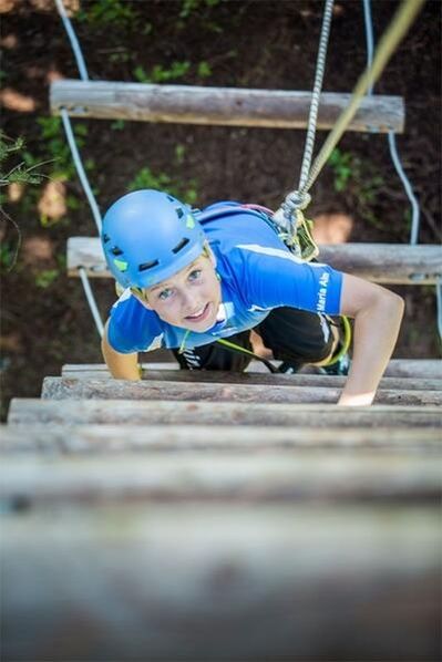 Eine Person mit einem blauen Helm klettert auf eine mit Seilen gesicherte Holzleiter